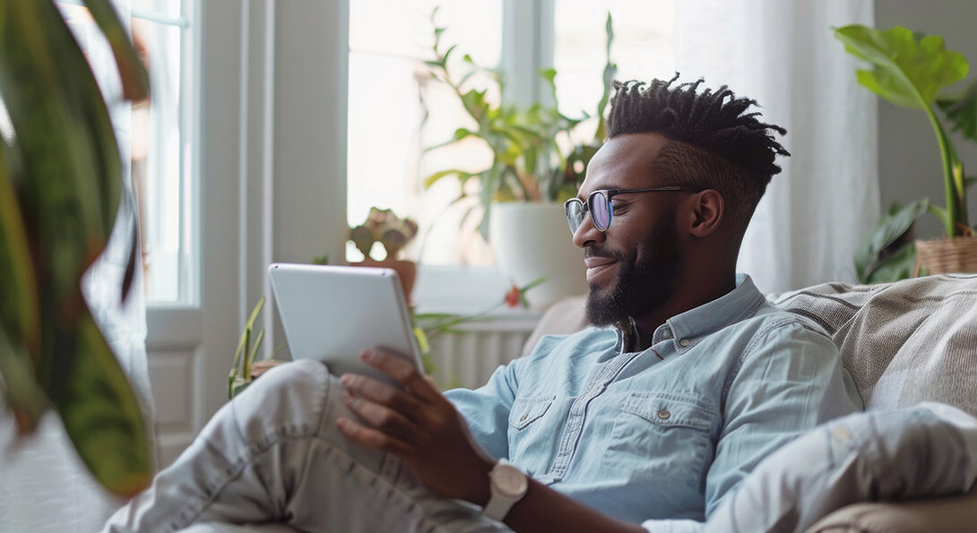 Person with dark skin, glasses, and beard looking at a tablet device. Their arm is holding the tablet while resting on a crossed knee. There are plants in the background and light coming in the window.