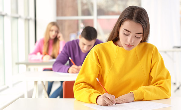 Three students taking an exam. Each one is in a bright color. They are handwriting the exam with red pencils.