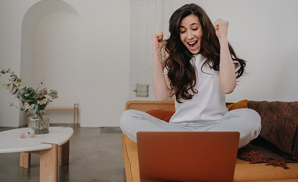 A person with long, dark hair sitting cross legged on a sofa. Their arms are up in a gesture of success or triumph. A laptop is open in front of them.